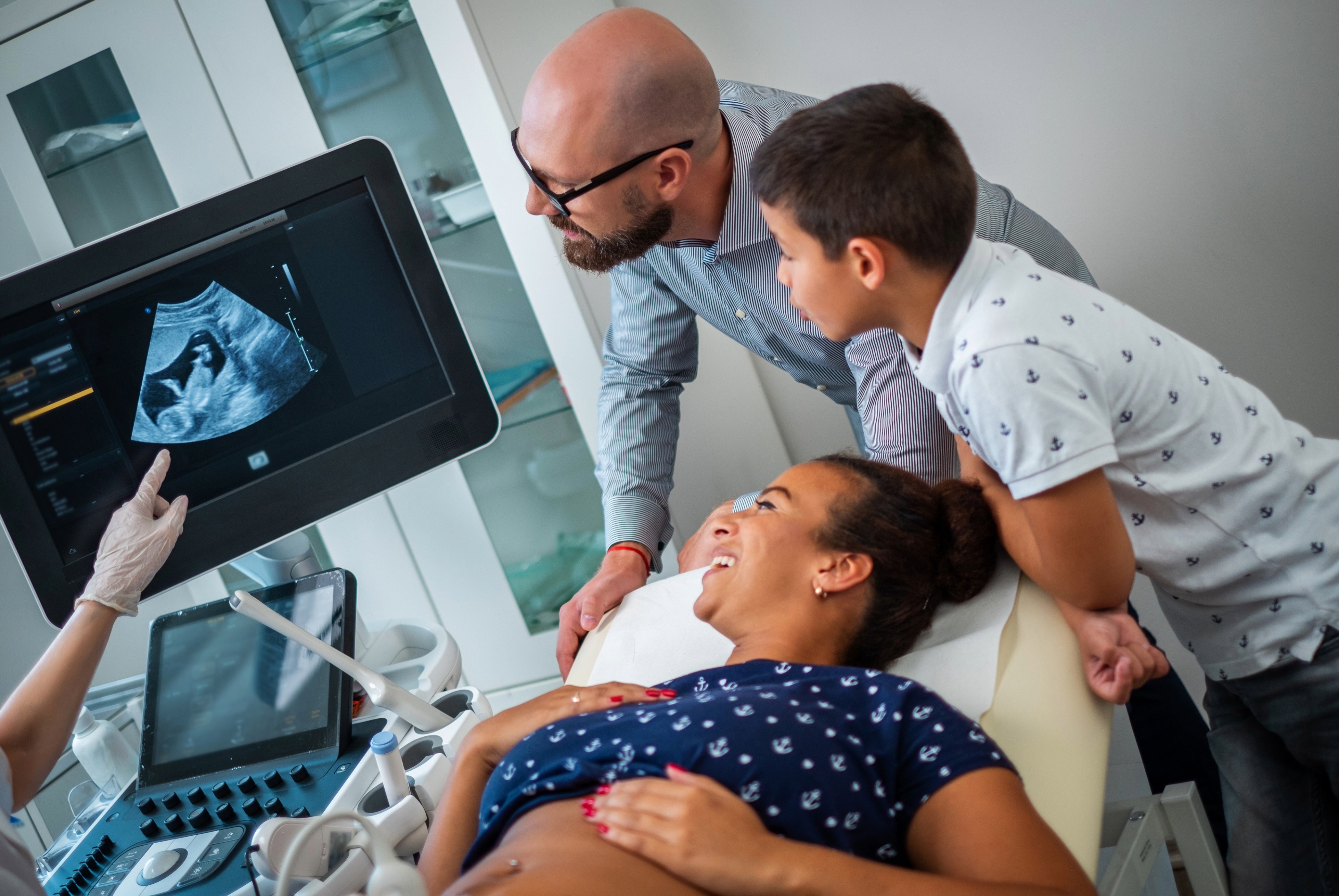 Una donna incinta è sottoposta ad ecografia ed insieme a compagno e figlio osserva l'immagine del nascituro sullo schermo.